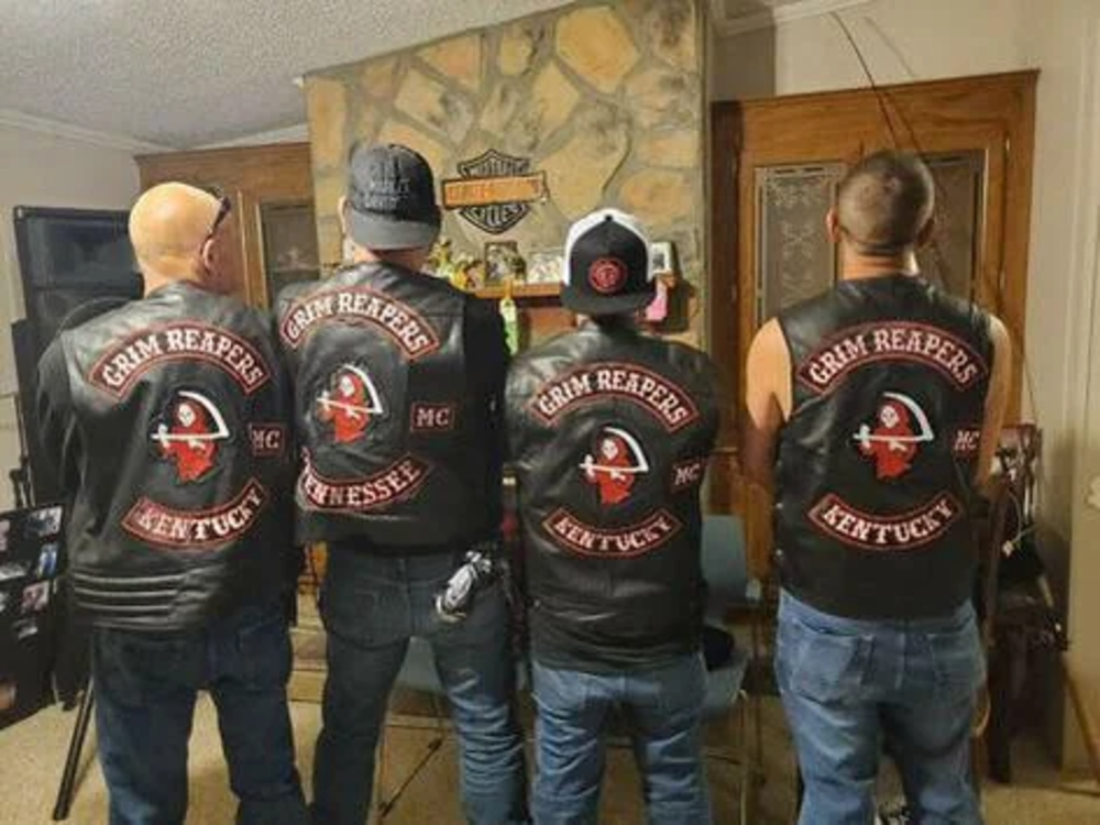 Four people stand indoors with their backs to the camera, wearing black leather vests with "Grim Reapers MC" and locations such as Kentucky and Tennessee. The vests display a winged skull logo. A wall with a Harley-Davidson emblem is in the background.