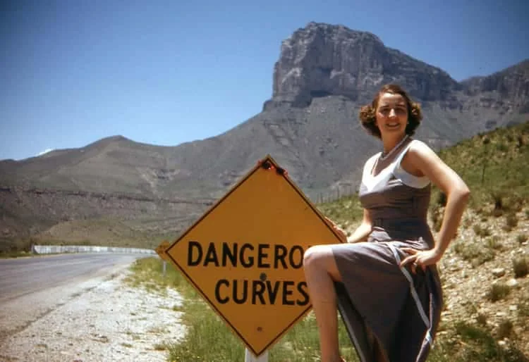 A woman in a gray sleeveless dress playfully poses with one leg raised on a roadside sign that reads "DANGEROUS CURVES." The background features a winding road and a mountainous landscape under a clear blue sky.