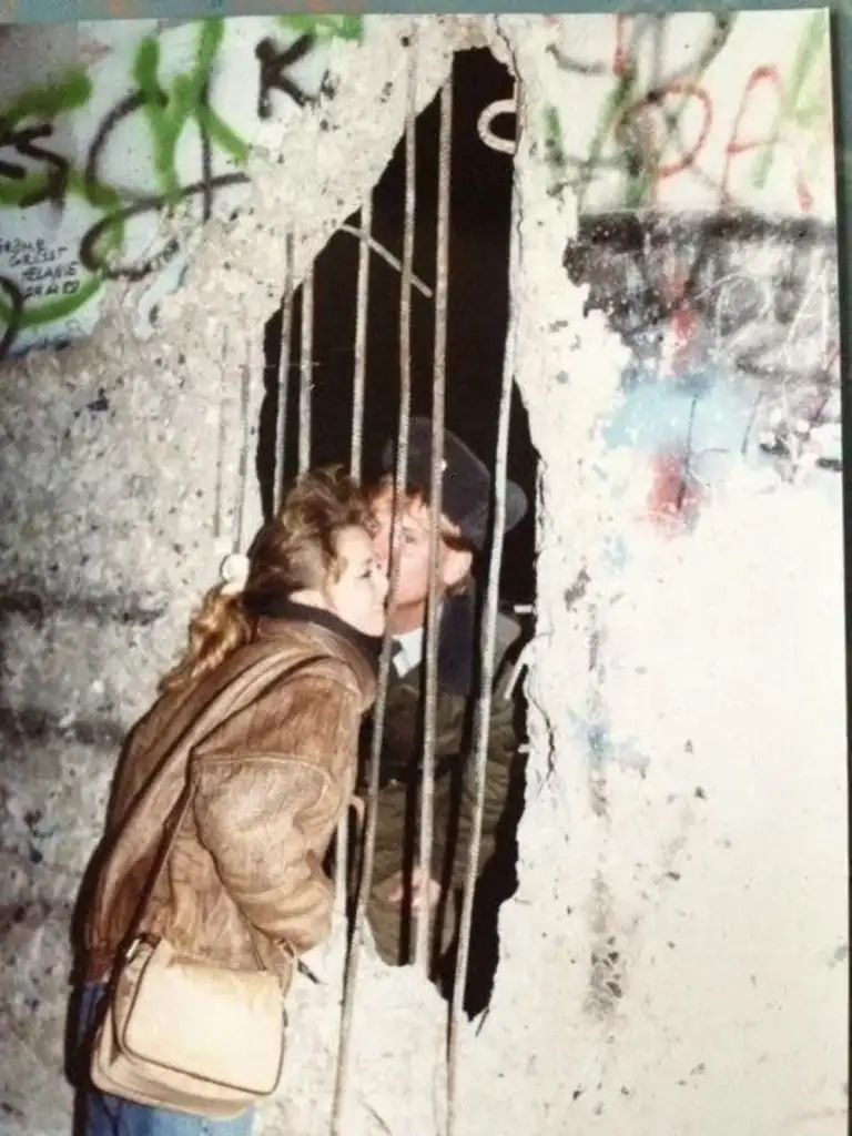 A woman and a man kiss through a gap in a graffiti-covered wall. Iron bars partially obstruct them. The woman carries a shoulder bag, and both wear jackets. The setting suggests a historic or significant location.