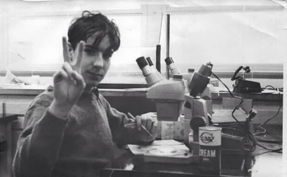 A person is sitting at a desk with a microscope, holding up a peace sign. The workspace has various items, including a container labeled "Dream." The setting appears to be a laboratory or study area with windows in the background.