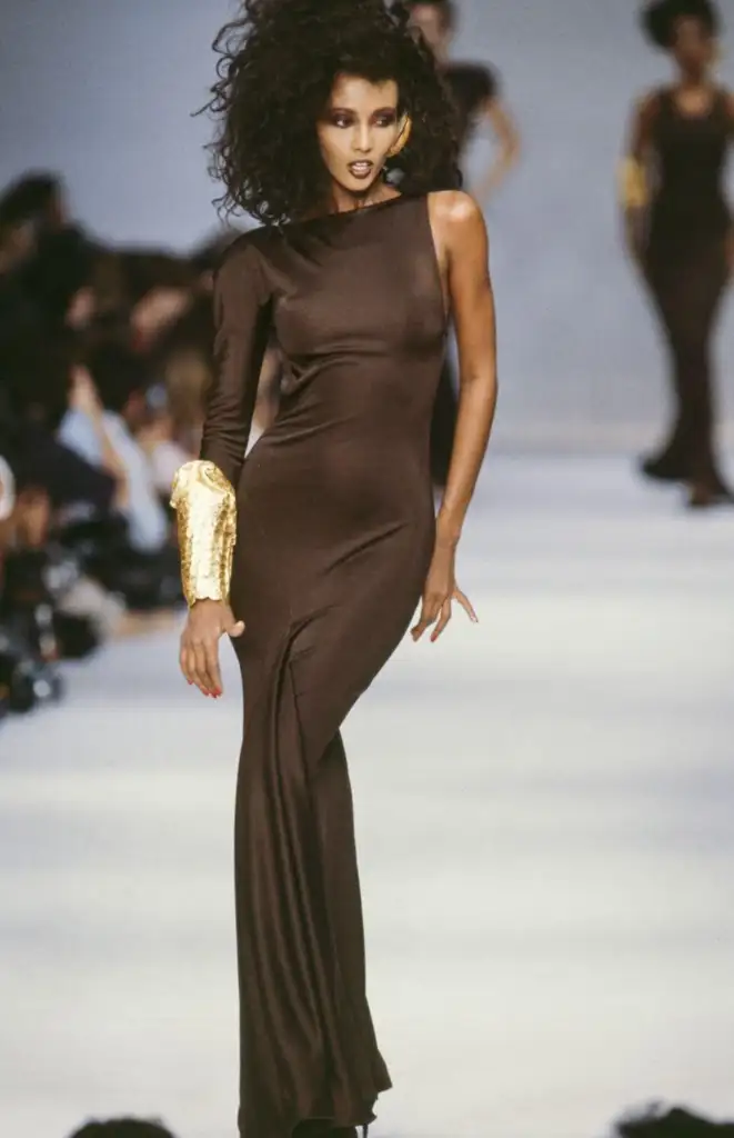 A model walks down the runway in a sleek, asymmetrical brown gown with one long sleeve and a gold accent near the wrist. Her voluminous curly hair frames her face, and she exudes confidence as she poses on the catwalk.