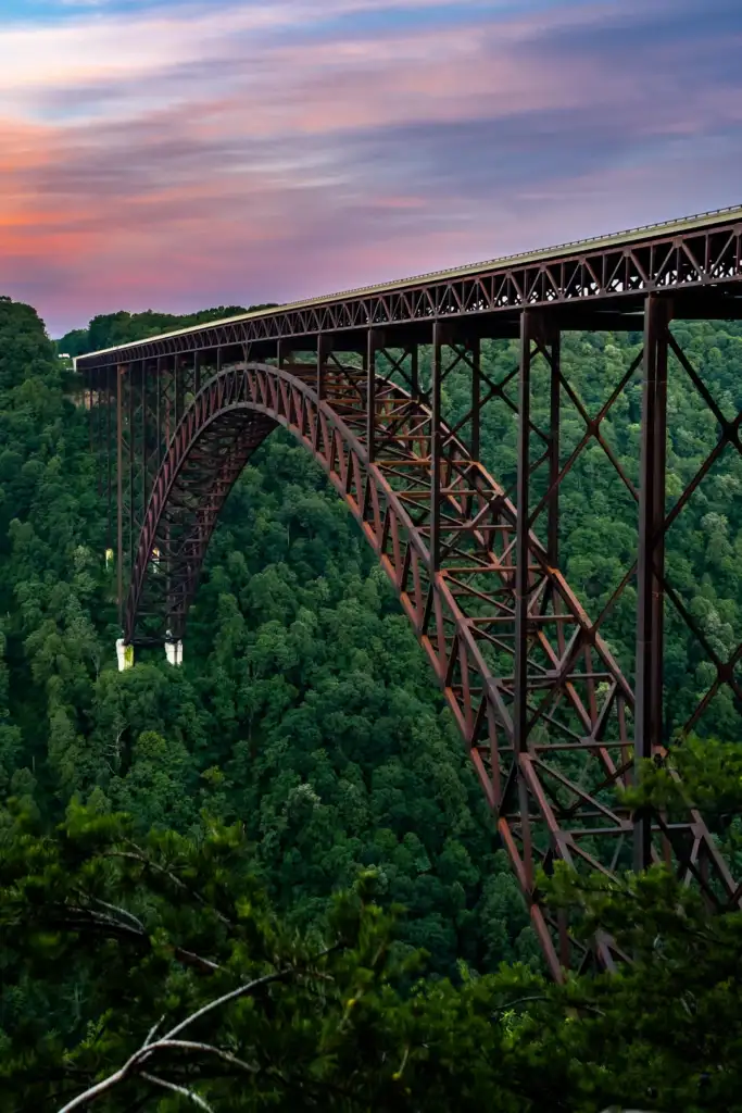 A large, arched steel bridge spans across a lush, green forested valley. The sky is painted with hues of orange, pink, and purple, suggesting sunset or sunrise. The bridge's intricate latticework is visible against the vibrant landscape.