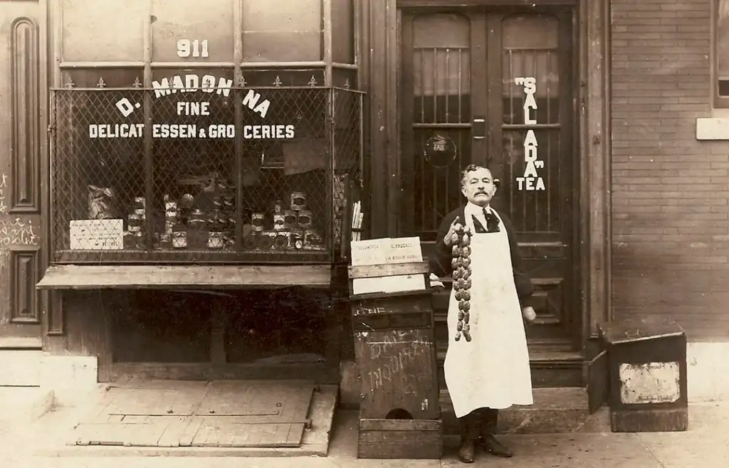 A man in a long apron stands holding a string of onions in front of a storefront displaying signs for delicatessen and groceries. The shop window has various goods, and the building number 911 is visible above the door.