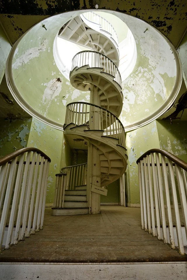 A spiraling wooden staircase ascends through a circular opening in a worn, peeling ceiling. The green walls and chipped paint reveal the structure's age. Two banisters flank the base of the staircase, leading upward in parallel curves.