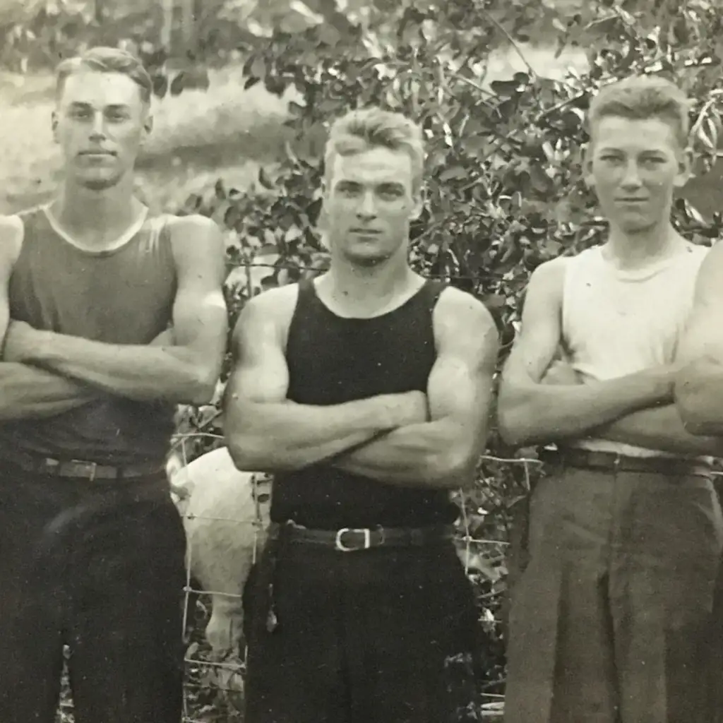 Three young men stand with arms crossed, wearing sleeveless tops, in front of a leafy background. They have short hairstyles and confident expressions, and the image appears to be a vintage black and white photograph.