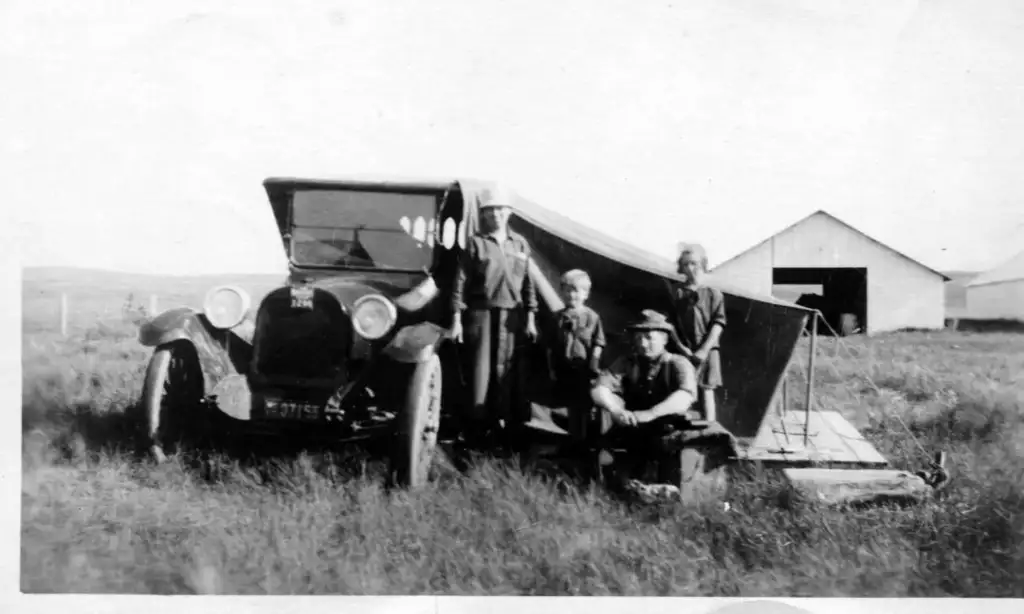 A black and white photo of four people and a dog in front of a vintage car with a makeshift tent attached. They are in a grassy field with a barn in the background. The scene suggests a roadside or camping setup from an earlier era.