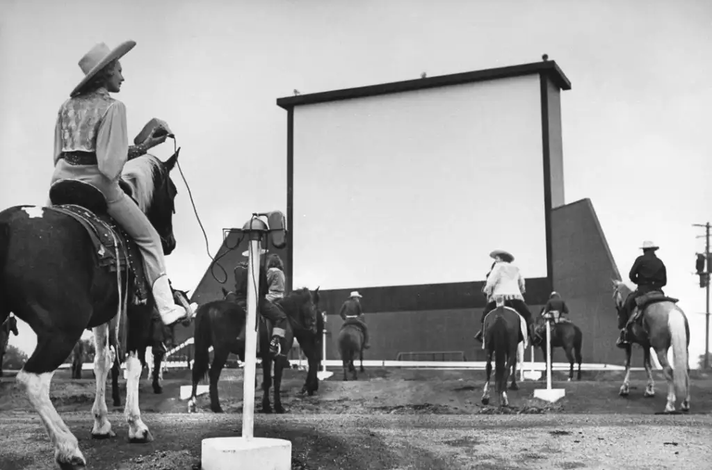 Six people on horses attend a drive-in cinema, facing a large blank screen. They are wearing cowboy hats and western attire. It's a black and white photo with an old-fashioned feel. The scene depicts a blend of tradition and modernity.