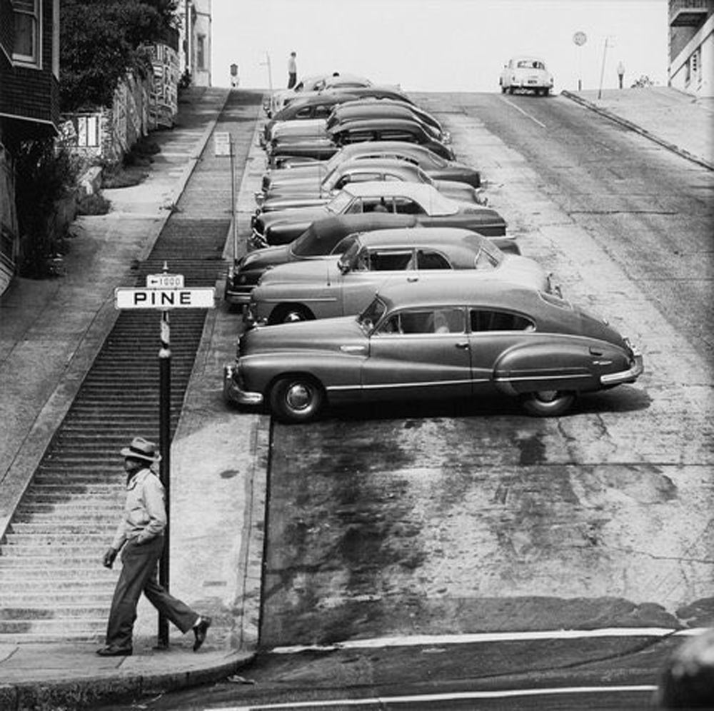 A man walks past a steep San Francisco street where vintage cars are parked perpendicular to the sloped road. A street sign reads "Pine." The street appears quiet with buildings alongside.