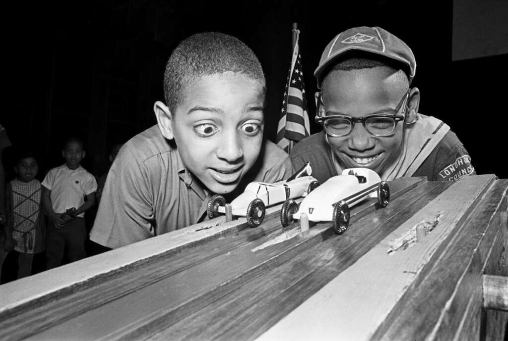 Two boys excitedly watch toy cars racing on a wooden track. One boy has wide eyes, and the other is smiling, wearing glasses and a cap. An American flag is visible in the background.