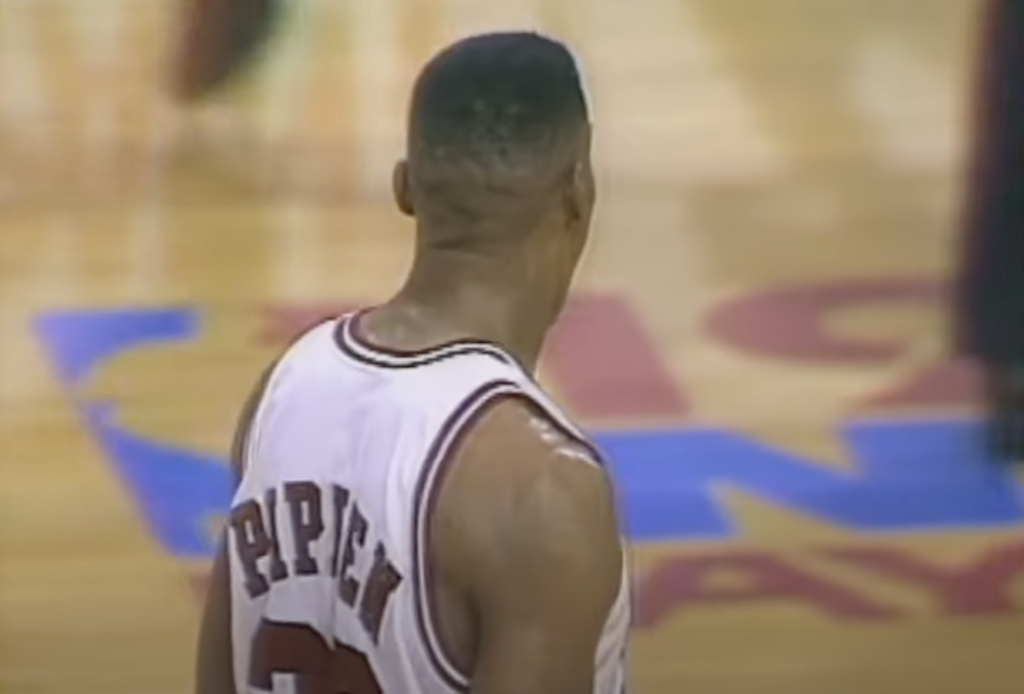 Basketball player wearing a white jersey with the name "Pippen" and the number "33" on the back, standing on a wooden court. The background shows a blurred logo on the floor.