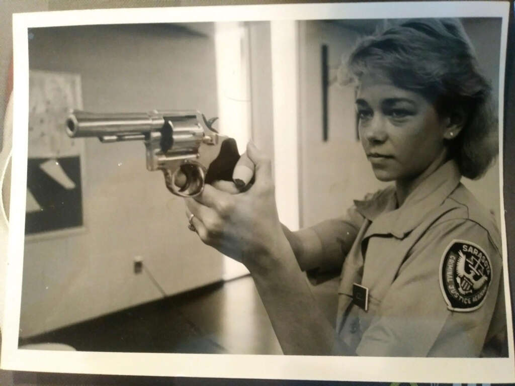 Black and white photo of a person in a uniform holding a revolver. They are aiming the gun with a focused expression, in an indoor setting. A patch on the sleeve reads "Sabra" and "Security Police.