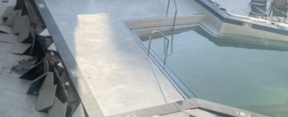 An outdoor hotel pool is partially drained, revealing cracked and collapsed tiles surrounding the water. Several lounge chairs are visible in the background on the pool deck.