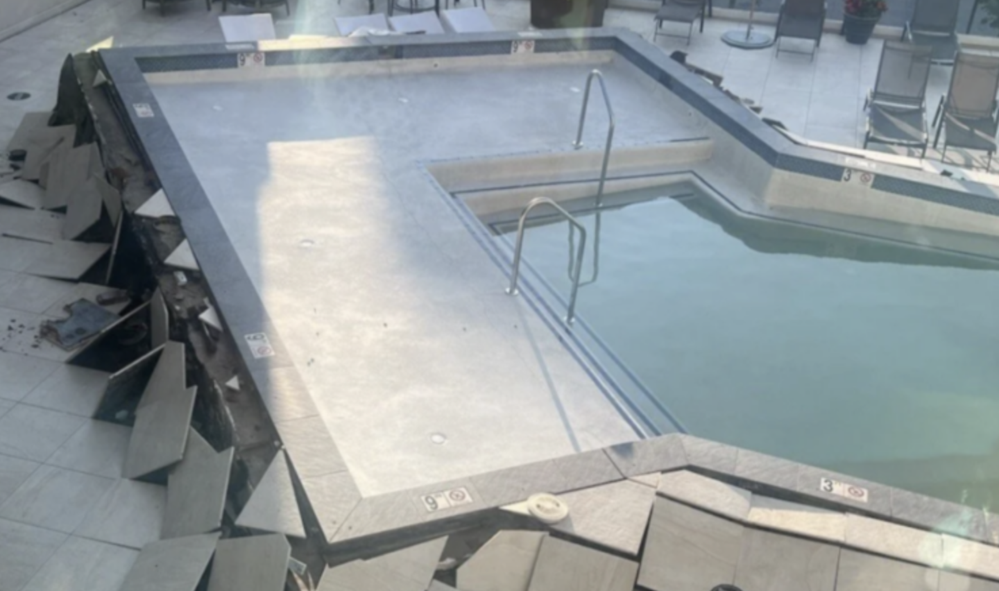 An outdoor hotel pool is partially drained, revealing cracked and collapsed tiles surrounding the water. Several lounge chairs are visible in the background on the pool deck.