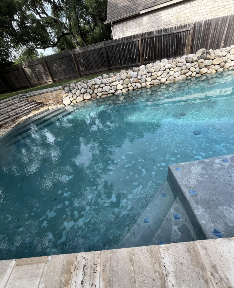 A backyard pool with clear blue water, bordered by stone steps and a rock wall. Trees and a wooden fence surround the area, and a stone-built house is partially visible in the background.