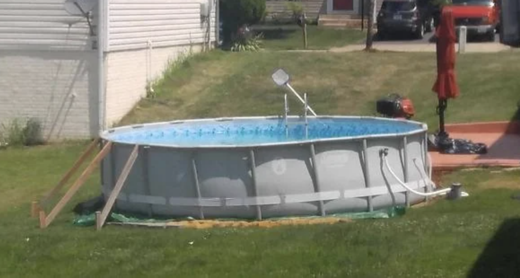 Above-ground pool in a backyard, surrounded by a grassy lawn. The pool is filled with water and has a small ladder for entry. A nearby patio area features a red umbrella. Homes and a driveway are visible in the background.