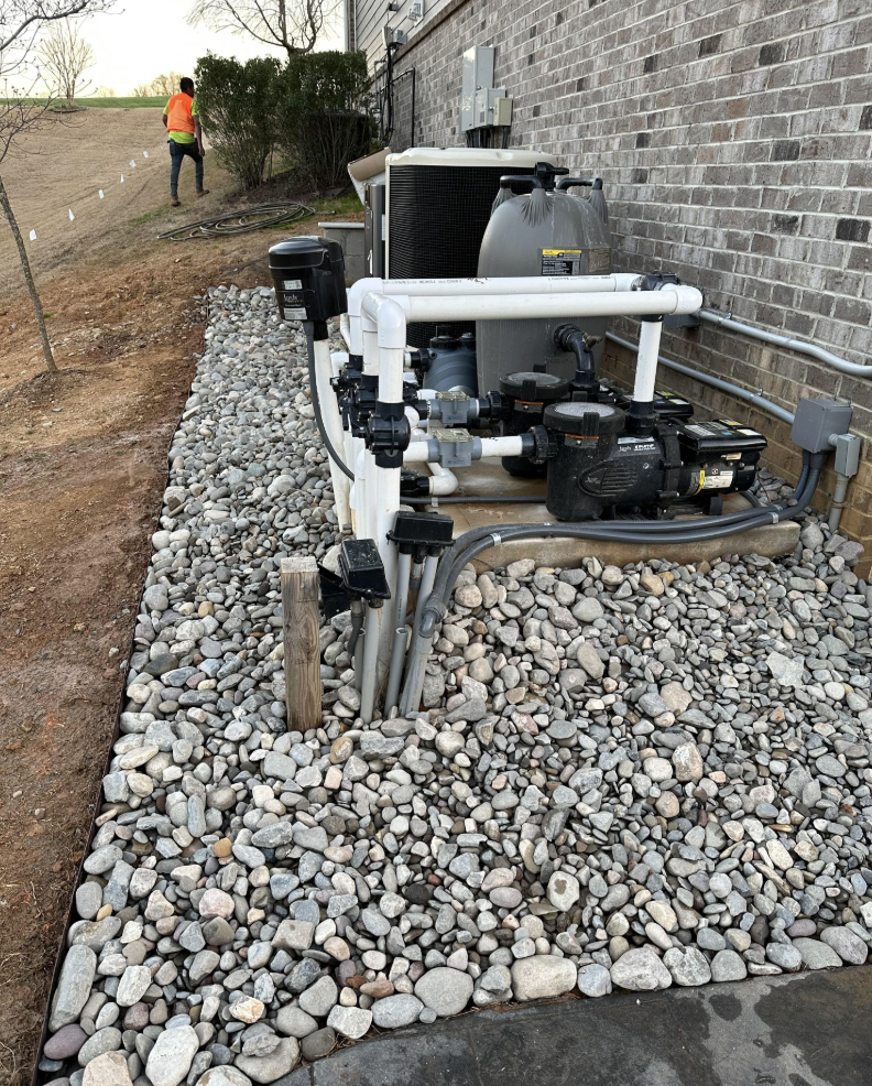 Pool equipment, including pipes and pumps, is installed on a bed of rocks next to a brick building. In the background, a person in an orange vest is walking on a dirt path near trees and grass.