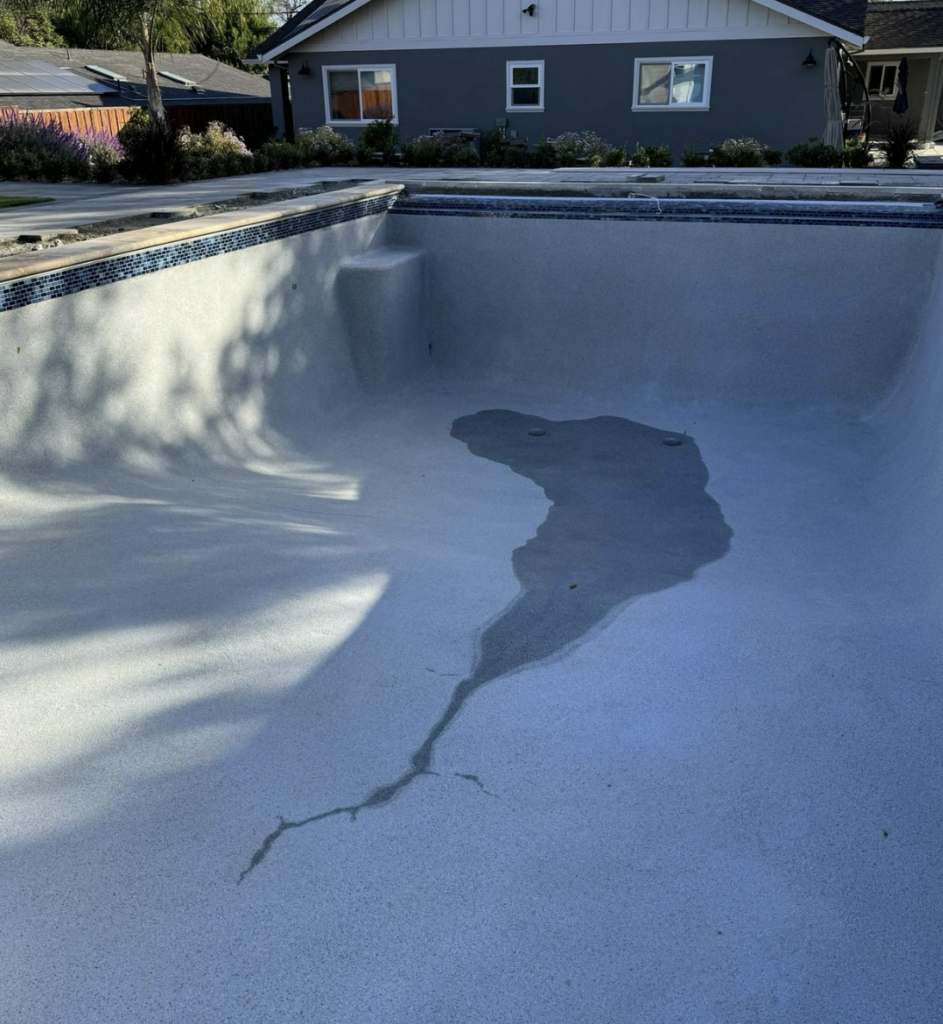 An empty swimming pool with a small, South America-shaped water puddle at the bottom. A house and trees are visible in the background. The pool's surface is light gray with shadow patterns.