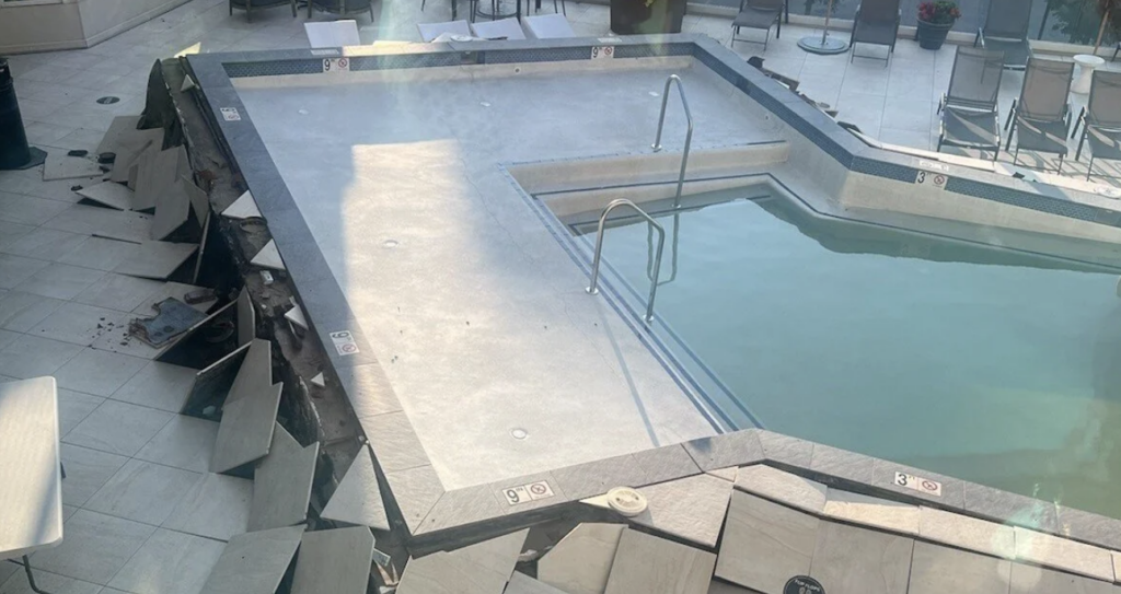 Damaged swimming pool with cracked tiles surrounding a sunken area. Water partially fills the pool. Patio chairs and potted plants are visible in the background.