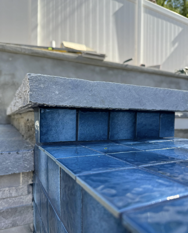 Swimming pool fails photo of close-up pic of a stone wall and steps with blue tiles on the side, partially wet from water. The background shows a blurred white fence and some construction materials, indicating ongoing work or renovation.