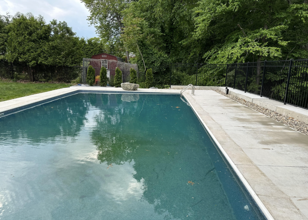 A rectangular outdoor swimming pool surrounded by a stone deck is shown. The pool is clear, reflecting nearby trees and the sky. Green trees line the background, and a red shed is visible at the far end of the pool area.