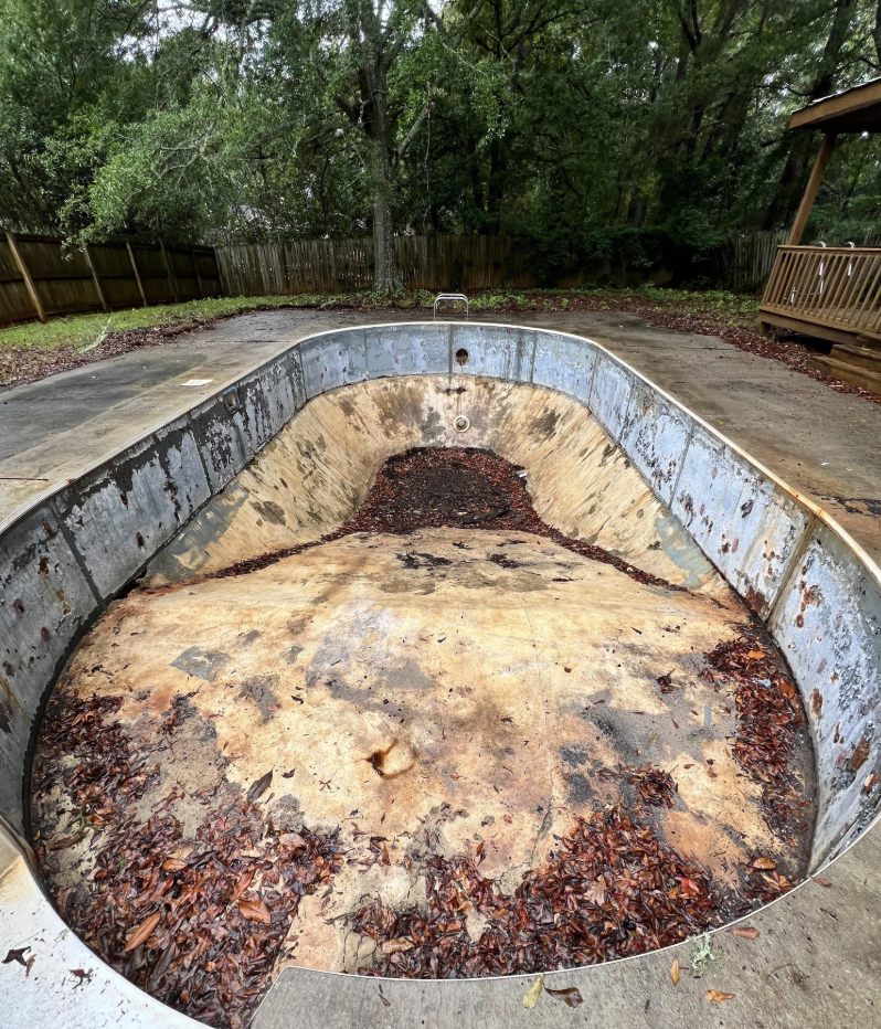 An empty, neglected swimming pool with rusted metal walls and a cracked, dirty floor filled with scattered dry leaves. Surrounding the pool is a concrete deck, and beyond it, a wooden fence and lush green trees are visible.