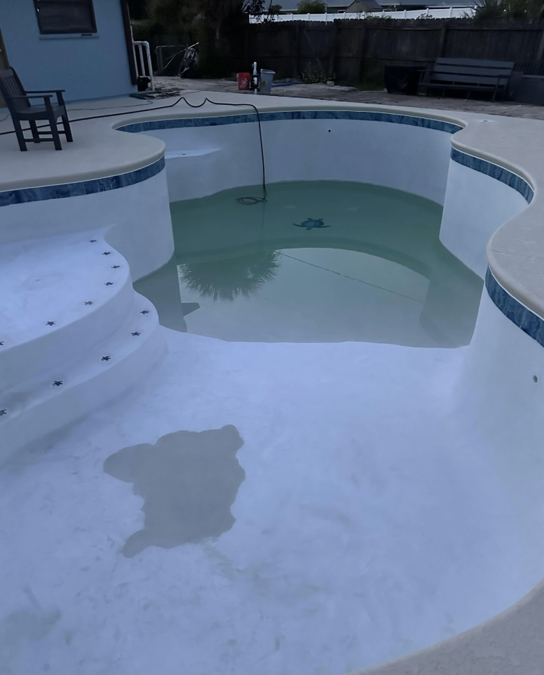 An empty swimming pool partially filled with water shows curved steps leading into it. The pool has a slight reflection of trees in the water, and a house and seating area are visible in the background.