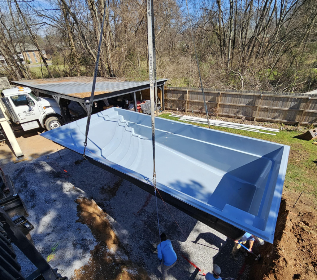 A crane is lowering a large, empty fiberglass pool into a prepared ground space in a backyard. Workers are guiding it in place. Surrounding the area are a truck, a shed, and trees without leaves.