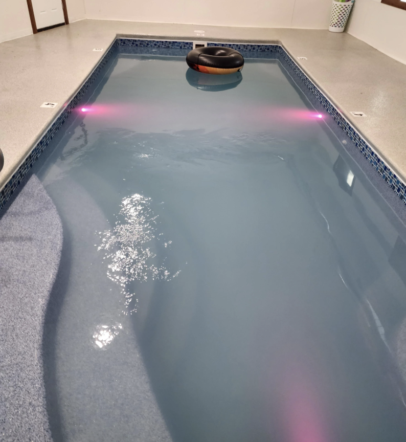 An indoor swimming pool with grey-blue tiles and pink lighting reflects on the water's surface. A black inflatable ring floats near one end. The pool is bordered by a smooth, light-colored deck.