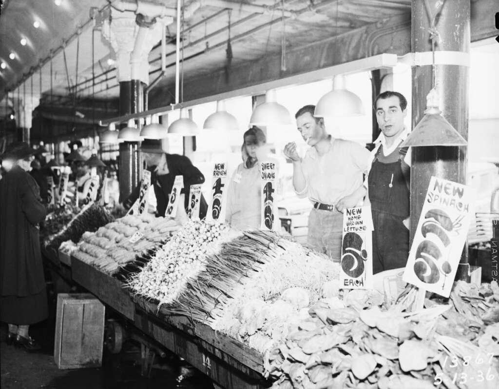 Black and white image of a bustling indoor market. Vendors stand behind a counter displaying various vegetables like lettuce, spinach, and peas. Price signs are visible. The market is well-lit with hanging lamps and has a lively atmosphere.