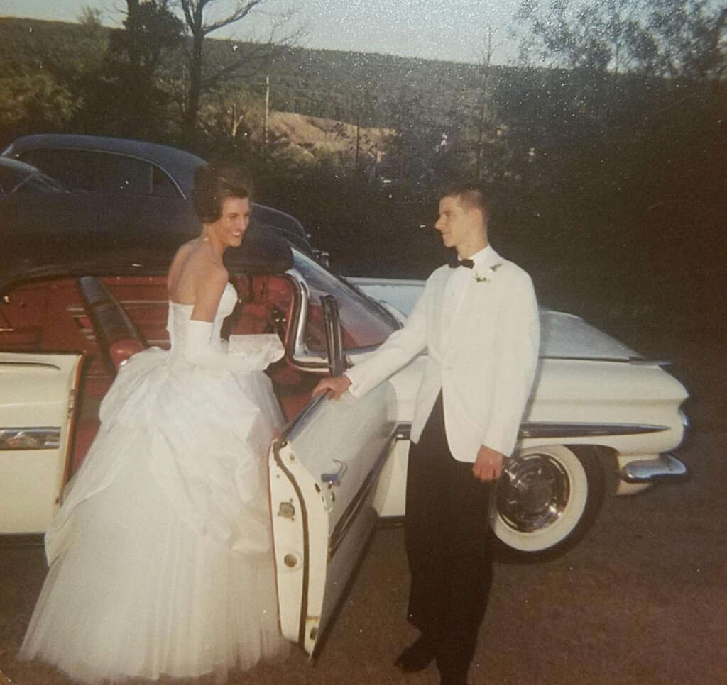 A woman in a white gown smiles while entering a classic car, held open by a man in a white tuxedo jacket. The car has red interior and is parked outdoors with trees and a hill in the background.