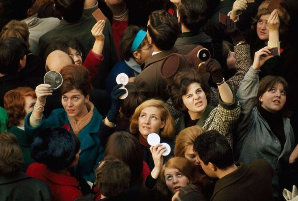 A crowd of people, mostly women, stand tightly packed together, holding compact mirrors upwards. They are dressed in mid-20th-century clothing. The individuals appear to be trying to catch a glimpse of something above them.