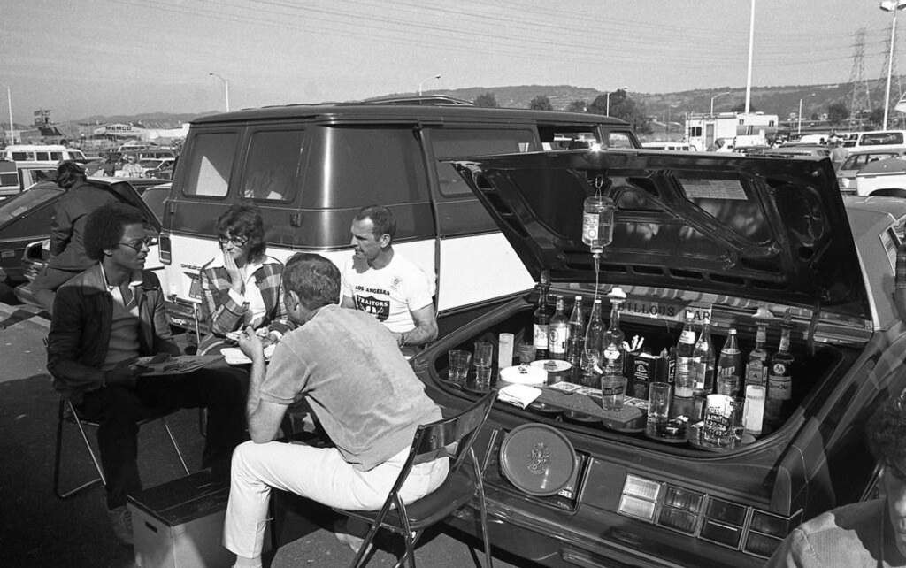 People are sitting on foldable chairs around a table in a parking lot. The trunk of a car is open, displaying various liquor bottles. There are vehicles in the background. The scene suggests a tailgate party.