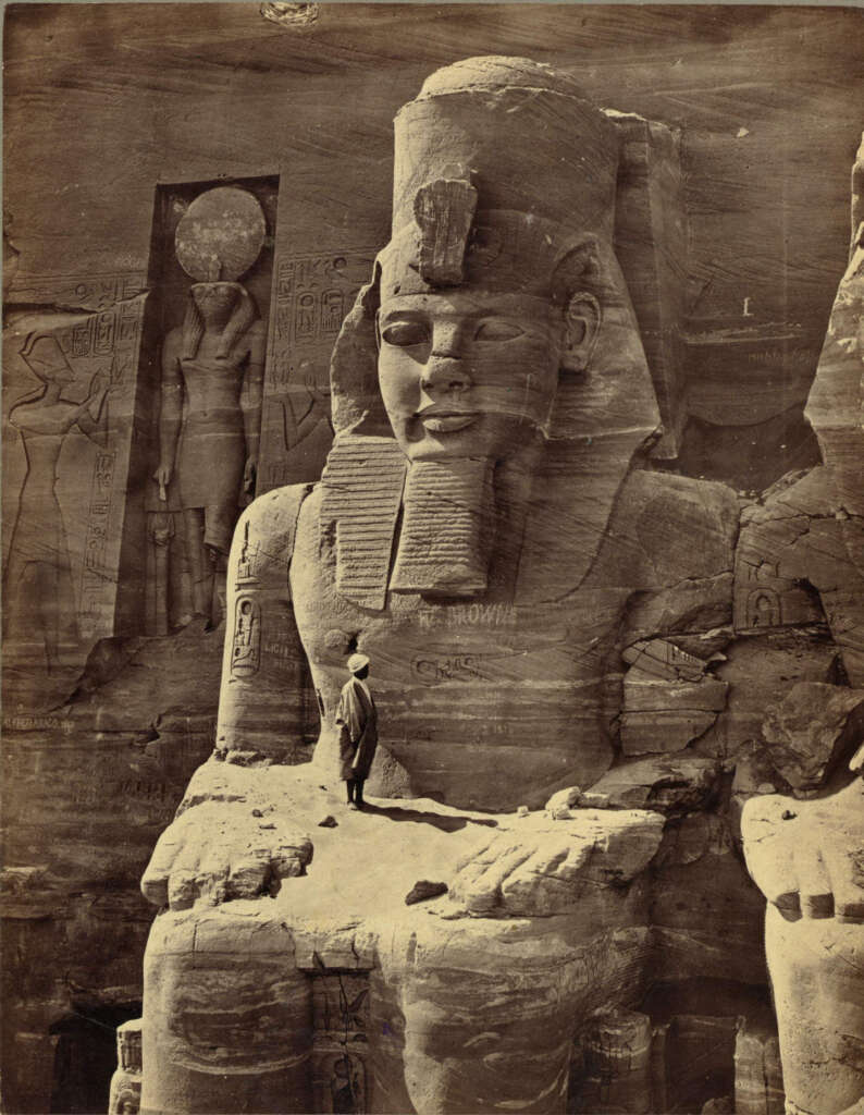 A person stands on the colossal stone foot of the Great Temple at Abu Simbel, Egypt. The massive seated statue of Pharaoh Ramses II dominates the scene, with intricate carvings and hieroglyphics visible in the background.