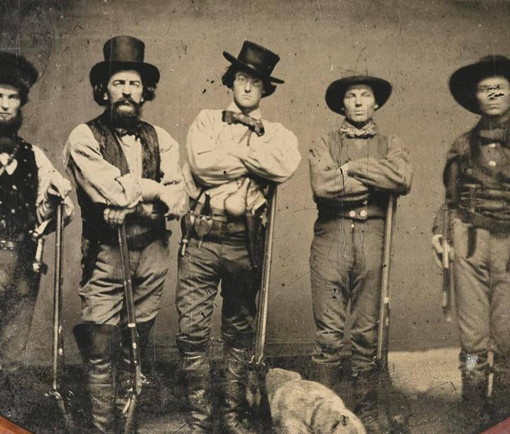 Five men in 19th-century attire stand with rifles, wearing hats and boots. They have stern expressions, and one man holds a pistol. A dog lies on the ground in front of them. The photo has a vintage, sepia-toned appearance.