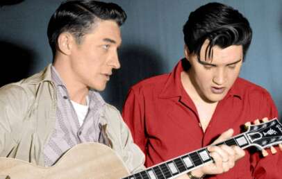 Two men are focused on a guitar. The man on the left, wearing a light-colored jacket, holds the guitar while the man on the right, in a red shirt, looks down. The background is a plain, dark color.