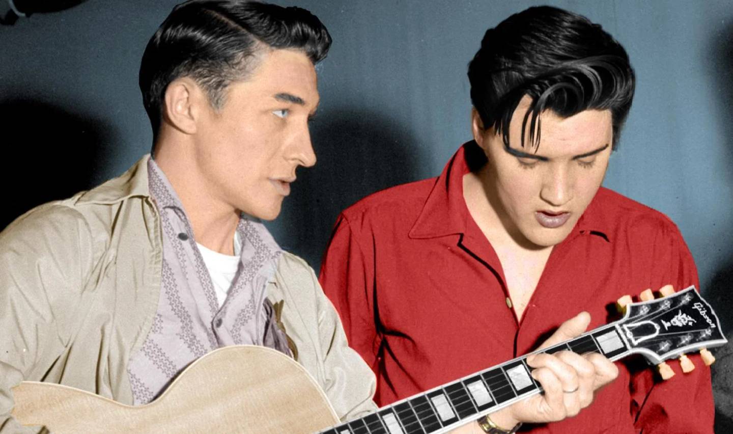 Two men are focused on a guitar. The man on the left, wearing a light-colored jacket, holds the guitar while the man on the right, in a red shirt, looks down. The background is a plain, dark color.