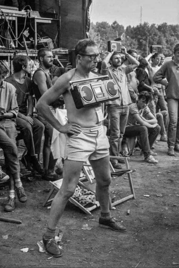 A man in shorts and glasses stands confidently with a large boombox strapped to his chest. He is outdoors, surrounded by a crowd of casually dressed people. Some are seated, others are standing. The scene appears to be at a lively outdoor event.