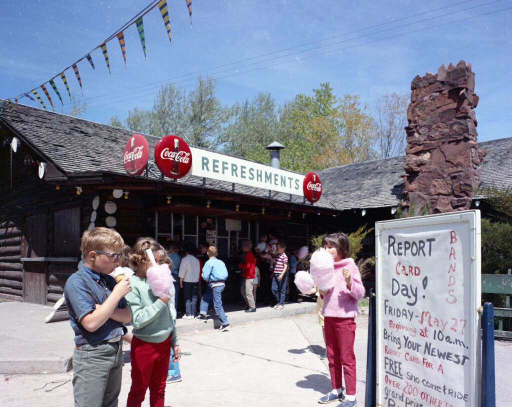 Children eating cotton candy near a rustic refreshment stand with Coca-Cola signage. A group of people is gathered in the background. A sign in the foreground advertises "Report Card Day" with various activities. Colorful flags are strung overhead.