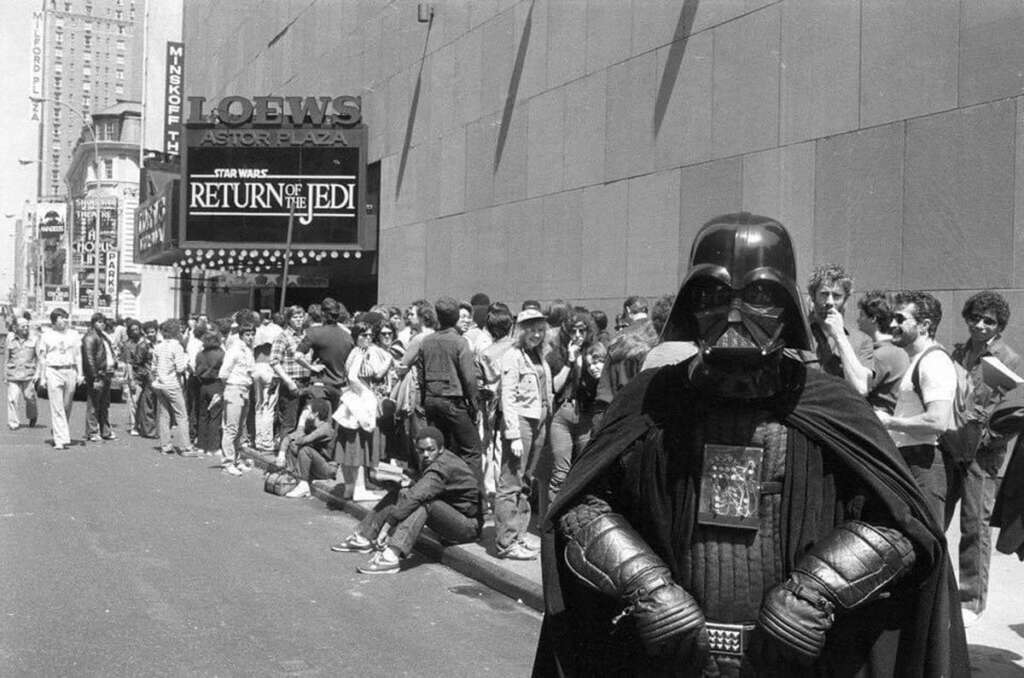 A person in a Darth Vader costume stands in front of a long line of people outside a theater. The marquee reads "Star Wars: Return of the Jedi." The scene captures a bustling city street with eager moviegoers.