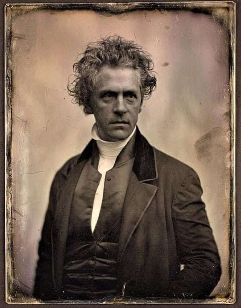 A sepia-toned daguerreotype of a man with curly hair, wearing a dark coat and waistcoat over a white shirt. He has a serious expression and is posed against a plain background, typical of 19th-century portraits.