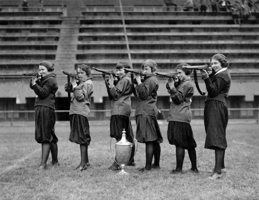 Six women in 1920s outfits stand in a line holding rifles, aiming to the left. A large trophy is placed on the ground in front of them. They are on a grassy field with empty stadium seating in the background.