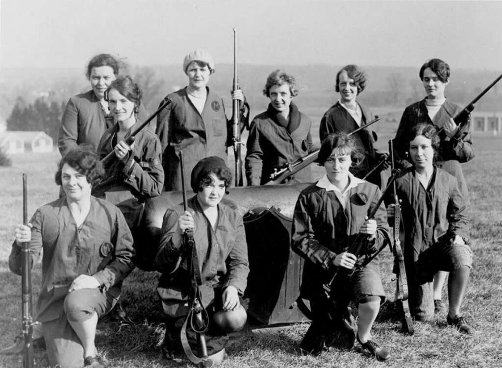 A group of vintage-clad women from the early 20th century pose outdoors with rifles. They are positioned around a barrel-like object, with some standing and others kneeling, all smiling. The background features a vast landscape under an open sky.