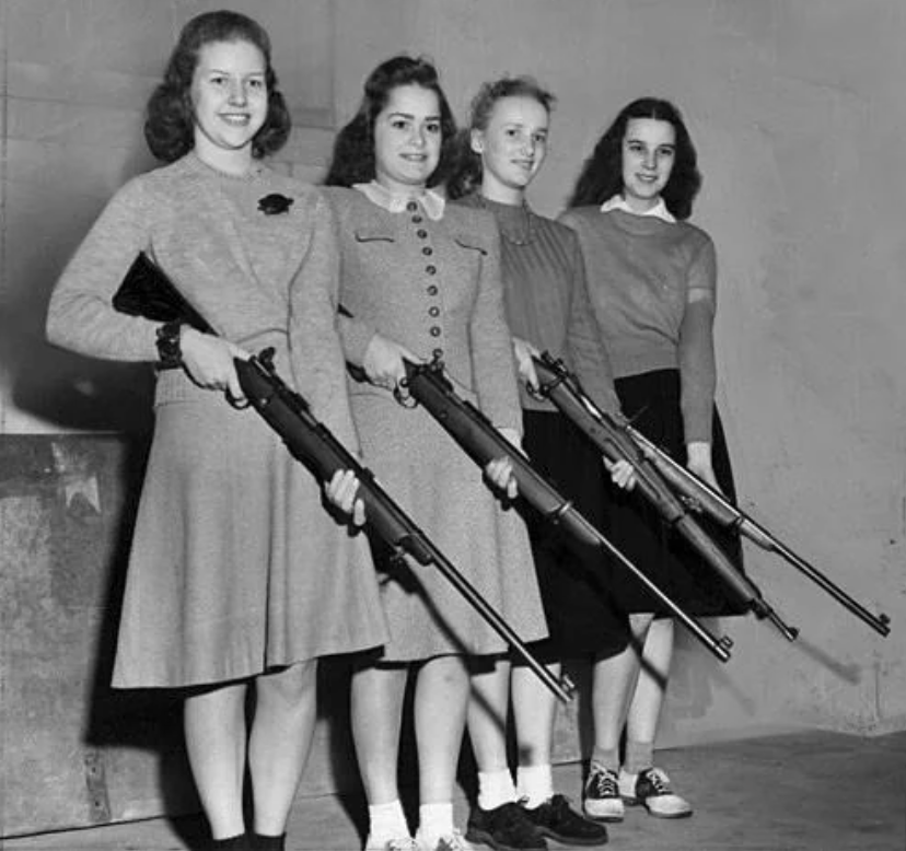 Four women stand in a row, each holding a rifle. They are dressed in mid-20th century attire, including skirts and cardigans. The background is plain, suggesting an indoor setting.