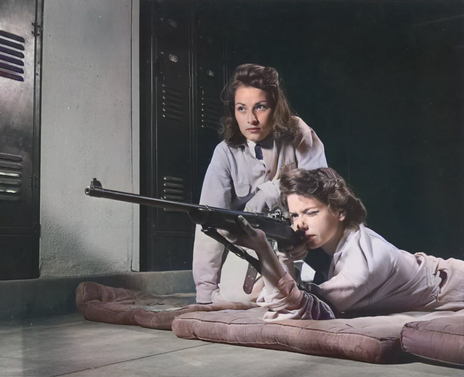 Two women in 1940s attire are in a dimly lit room. One woman is lying on mats, aiming a rifle, while the other kneels behind her, observing. Lockers are visible in the background.