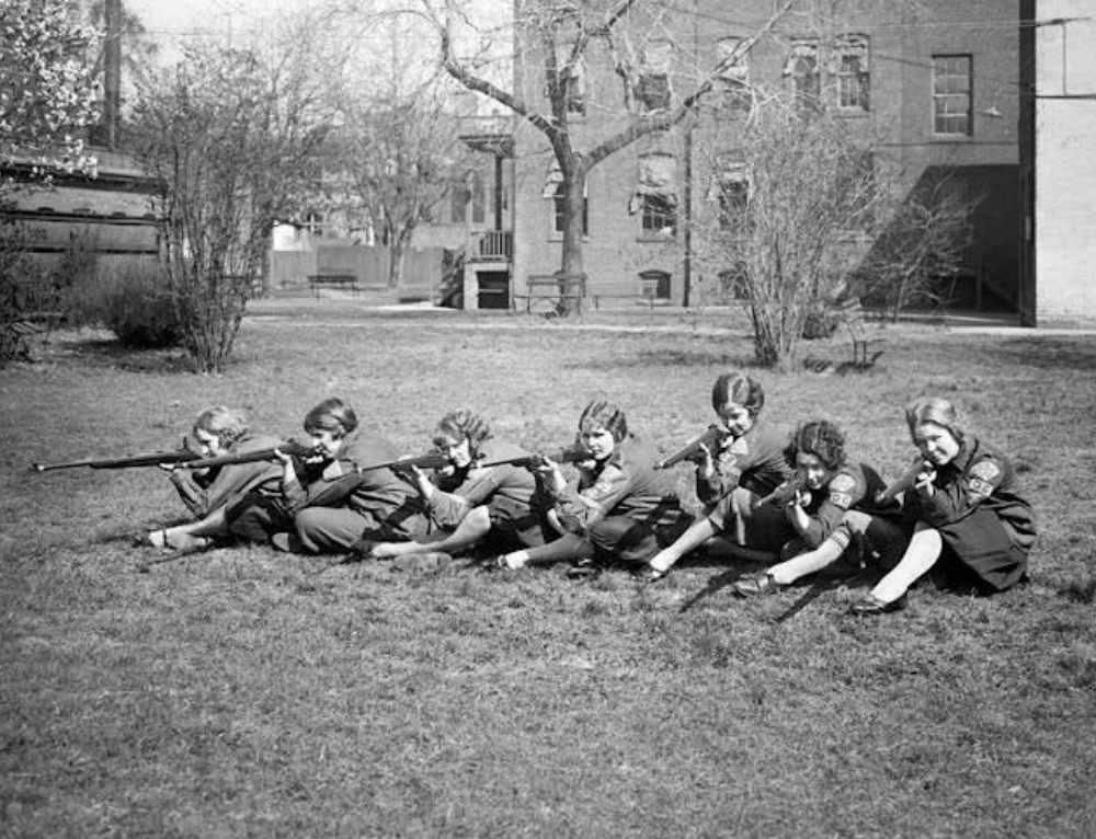 A group of six young women in uniforms sit in a row on grass, aiming rifles. They appear focused and intent. The background features trees and brick buildings. The scene is set in an outdoor, possibly educational or training environment.