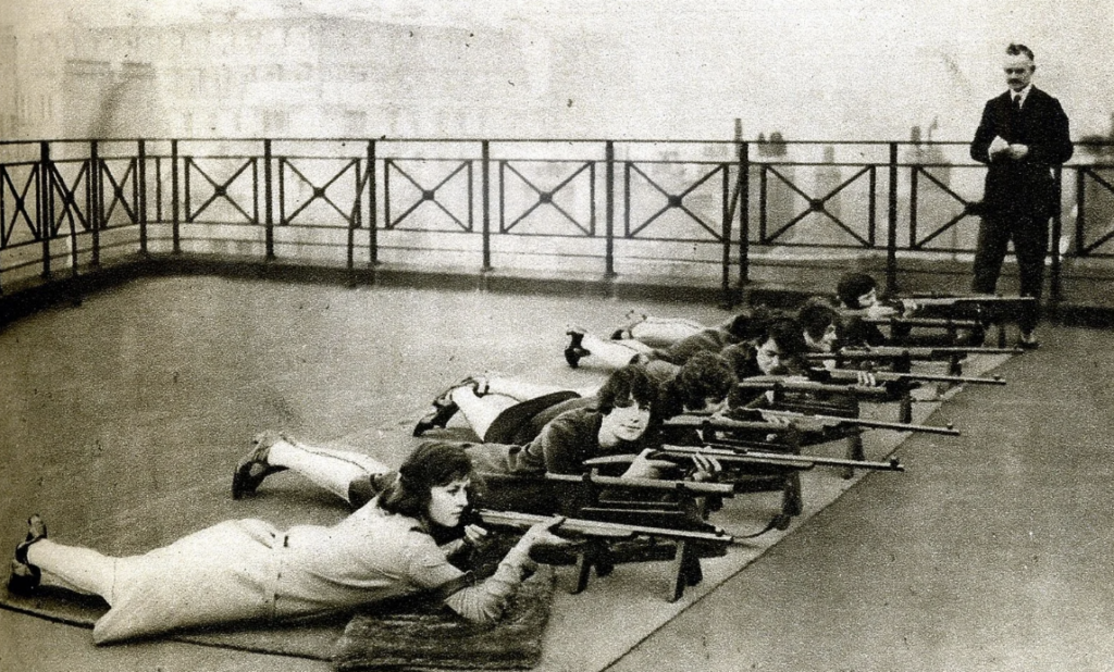 A historical black and white photo showing a group of women lying on their stomachs on a rooftop, each aiming a rifle. They are lined up next to each other. A man stands nearby observing them. The background features a hazy cityscape.