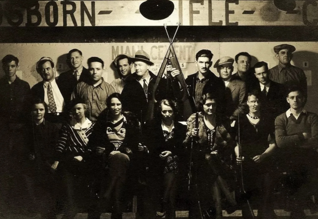 A black-and-white photo of a group of men and women in vintage clothing posing in front of a sign reading "MIAMI CEMENT." Two men in the center hold rifles crossed in front of them. The scene captures a historic, confident group ambiance.