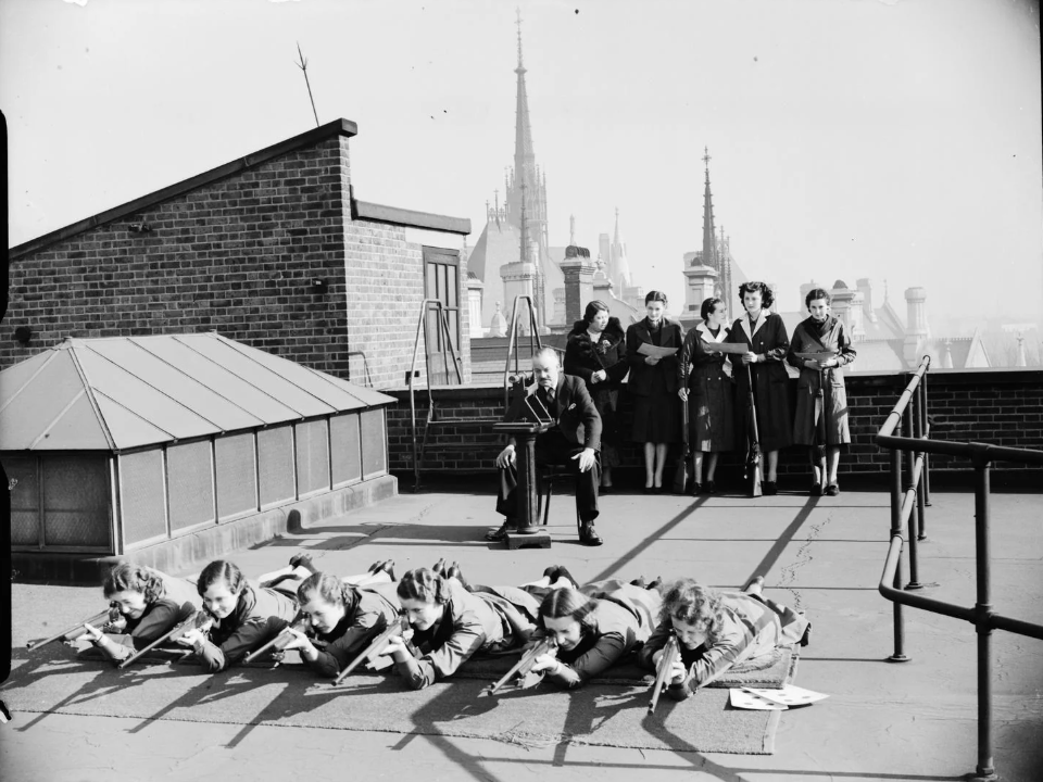 A group of people in 1940s attire stand and sit on a rooftop. A line of women lie on the ground aiming rifles, while two women and one man stand behind them, observing. A gothic-style building spire is visible in the background.