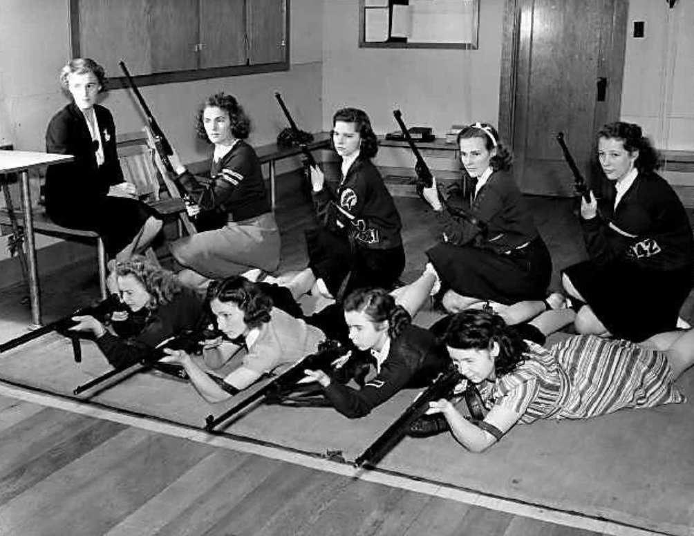 A vintage black-and-white photo shows a group of young women practicing rifle shooting indoors. Four women are seated, holding rifles, while four more lie prone on mats, aiming rifles. An instructor sits at a desk observing them.