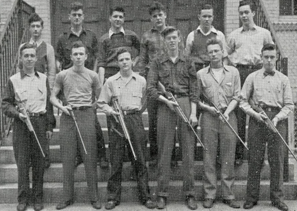 A black and white photo of thirteen men from a rifle club. Eight are standing in front holding rifles, and the remaining five are standing behind them on steps. The background features a brick building with doors.